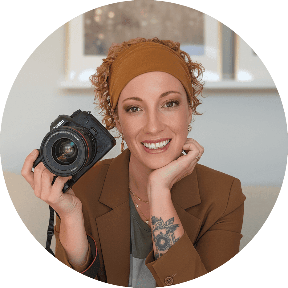 Une femme souriante aux cheveux bouclés et au bandeau tient un appareil photo, prête à prendre une photo. Elle porte un blazer brun et a un tatouage floral sur l'avant-bras. Assise à l'intérieur à Granby, elle appuie son menton sur sa main dans un cadre scolaire.