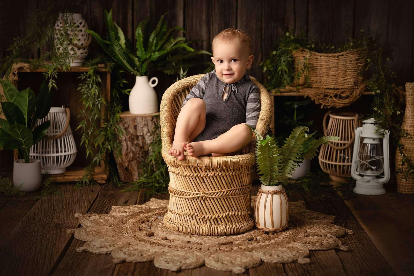 Service photo scolaire pour écoles et CPE 5 Un jeune enfant en tenue grise est assis les jambes croisées sur une chaise en osier, entouré de diverses plantes, de paniers et de décorations rustiques sur un sol en bois.