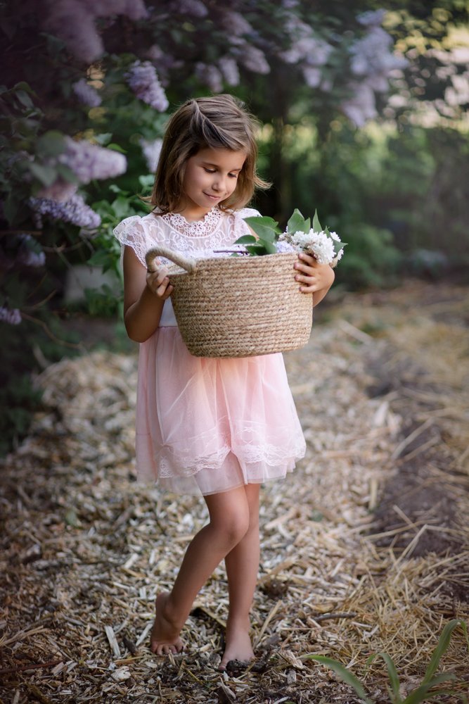 Une jeune fille vêtue d'une robe rose se tient pieds nus sur une allée de jardin, tenant un panier tressé rempli de fleurs, entourée de verdure et de buissons de lilas en fleurs.