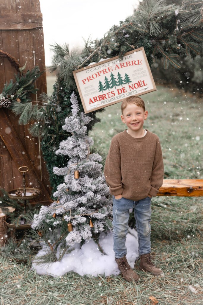 Un jeune garçon vêtu d'un pull marron et d'un jean sourit à côté d'un sapin de Noël artificiel enneigé, sous une arche rustique avec un panneau indiquant "Arbres de Noël", dans un cadre hivernal en plein air avec de la neige légère.