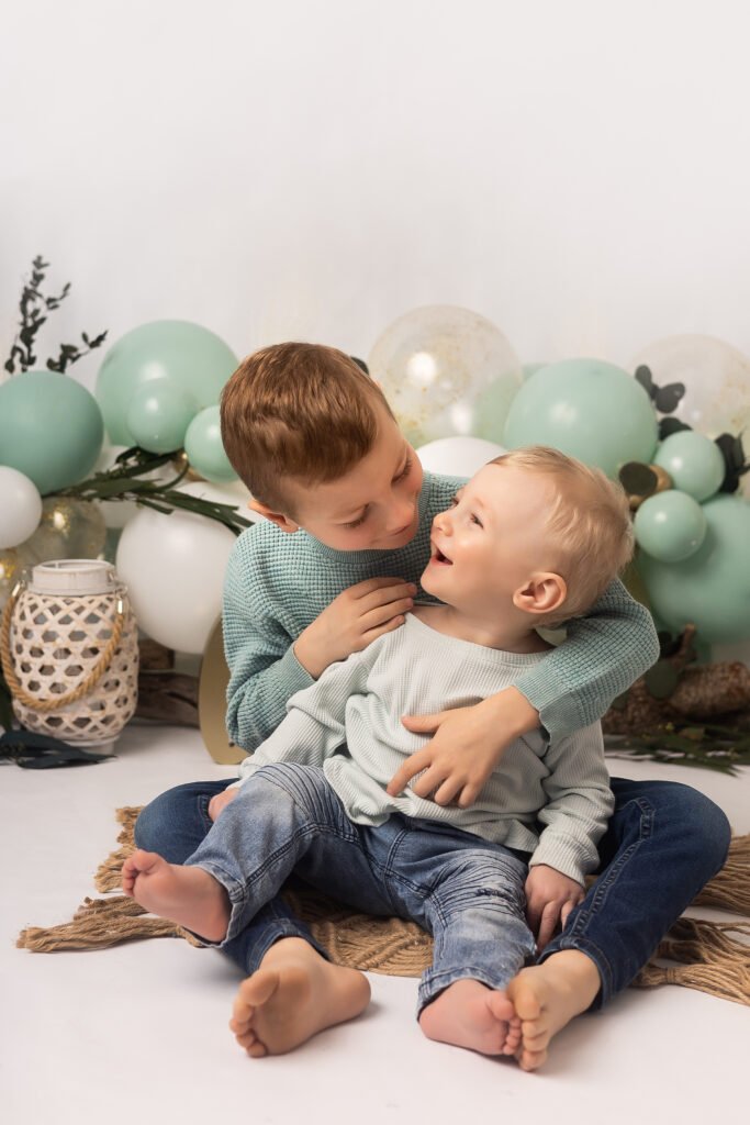 Deux jeunes garçons sont assis sur un tapis, souriant et se serrant l'un contre l'autre. Le plus âgé est assis derrière le plus jeune et l'entoure de ses bras. Des ballons verts et blancs et des objets décoratifs se trouvent à l'arrière-plan.