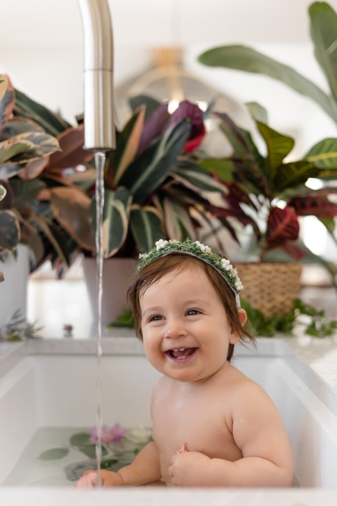 Un bébé souriant portant un bandeau floral est assis dans un évier blanc rempli d'eau et de pétales de fleurs, avec des plantes en arrière-plan et de l'eau qui coule du robinet.
