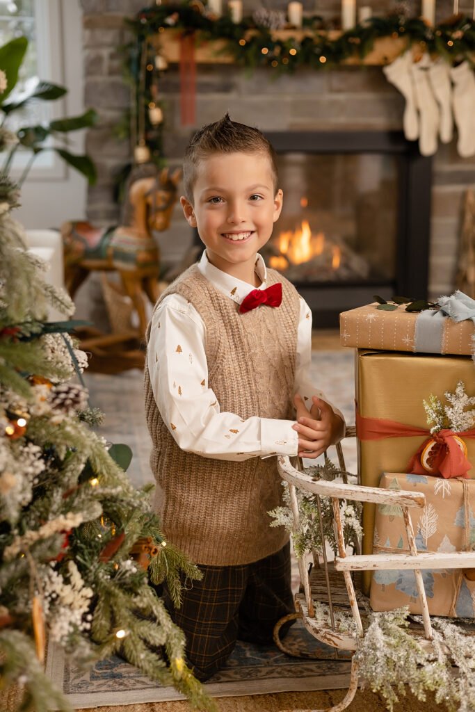 Un jeune garçon vêtu d'un gilet et d'un nœud papillon rouge est agenouillé près d'un traîneau contenant des cadeaux emballés, souriant devant un sapin de Noël décoré et une cheminée accueillante avec des bas et des décorations de vacances.