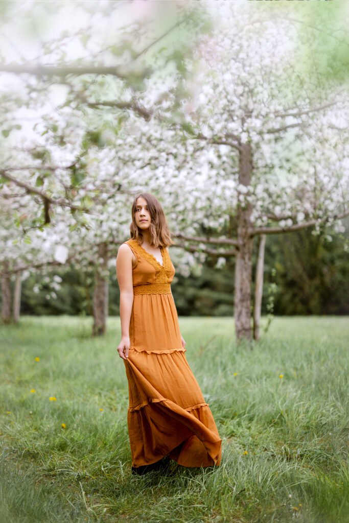 Une femme vêtue d'une longue robe orange rouille se tient debout sur l'herbe verte d'un verger aux arbres en fleurs, tenant sa robe et regardant de côté avec une expression calme.