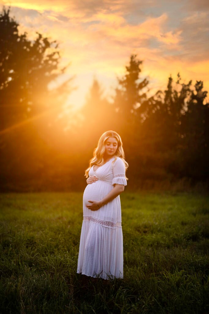 Une femme enceinte vêtue d'une longue robe blanche se tient dans un champ herbeux au coucher du soleil, berçant doucement son ventre. Le soleil se couche derrière elle, projetant une chaude lueur dorée et des silhouettes d'arbres en arrière-plan.