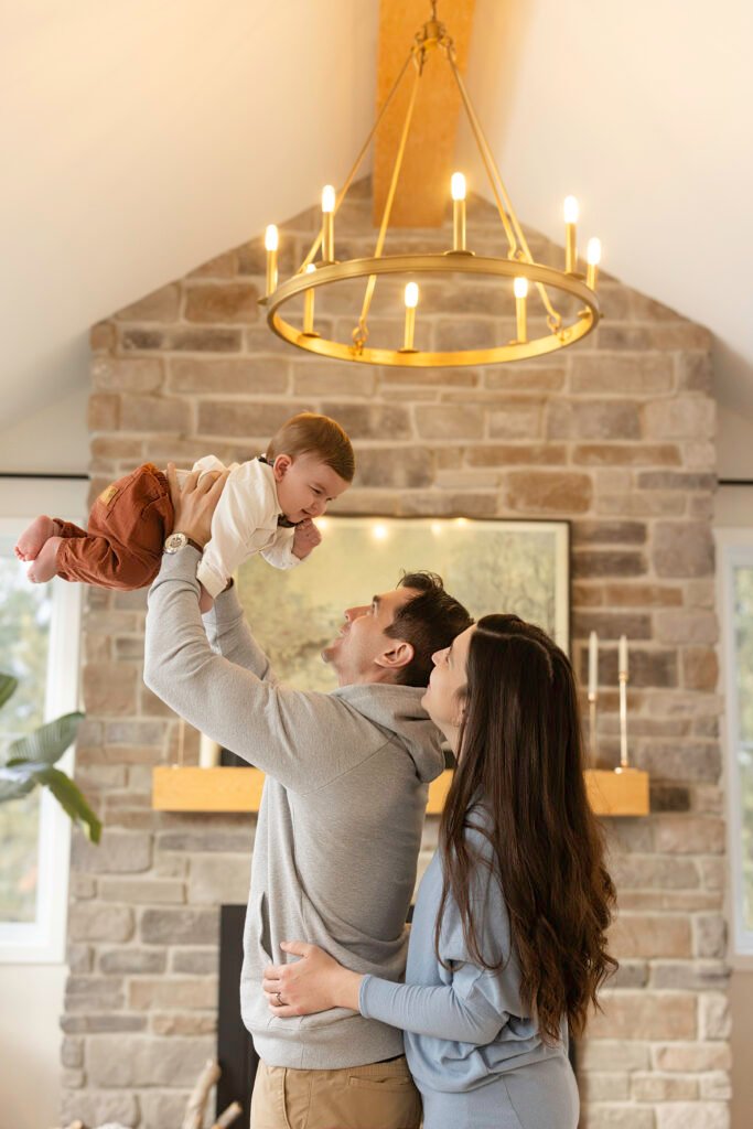 Un homme soulève un bébé souriant dans les airs tandis qu'une femme se tient à côté d'eux, les observant et souriant. Ils se trouvent dans un salon confortable avec une cheminée en pierre et un lustre doré au-dessus de leur tête.
