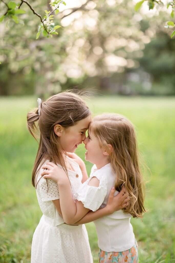 Deux jeunes filles se tiennent dans un champ herbeux, souriant et se touchant le front. Elles portent des vêtements clairs et semblent heureuses, la lumière douce du soleil illuminant la scène.