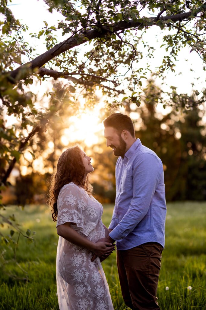 Un couple se tient dans un champ herbeux au coucher du soleil, se tenant par la main et se regardant l'un l'autre. La femme, qui porte une robe en dentelle, semble enceinte. La lumière du soleil filtre à travers les branches d'arbres au-dessus d'eux, créant une atmosphère chaude et lumineuse.