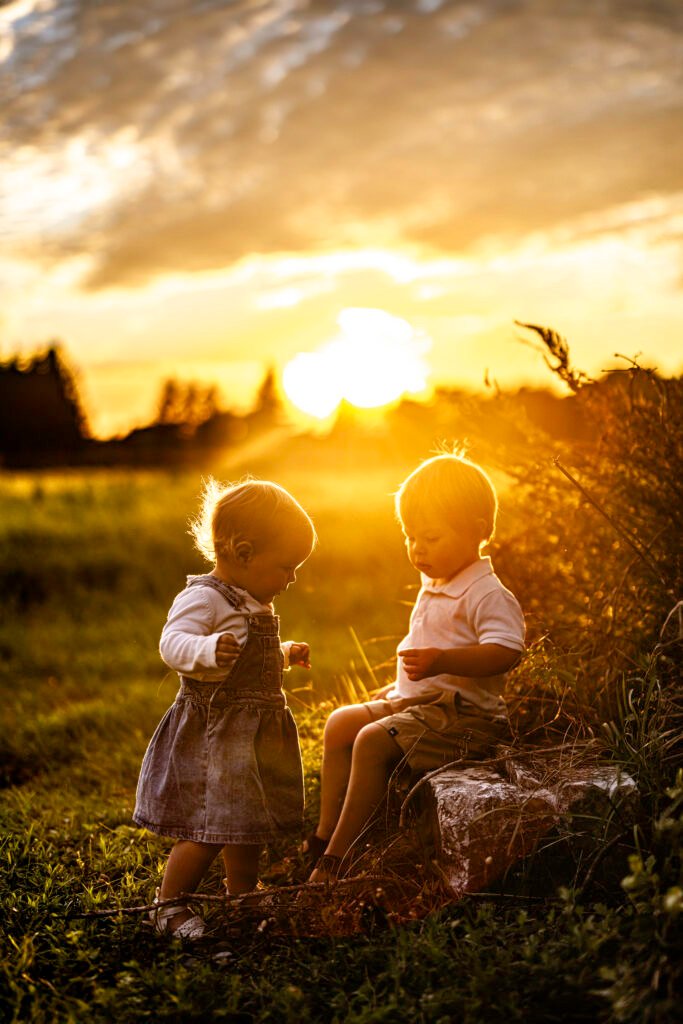 Deux jeunes enfants jouent ensemble en plein air au coucher du soleil, entourés d'herbe et de la lumière chaude et dorée du soleil. L'un d'eux porte une robe en jean, tandis que l'autre est assis sur un rocher. Tous deux semblent joyeux et engagés l'un envers l'autre.