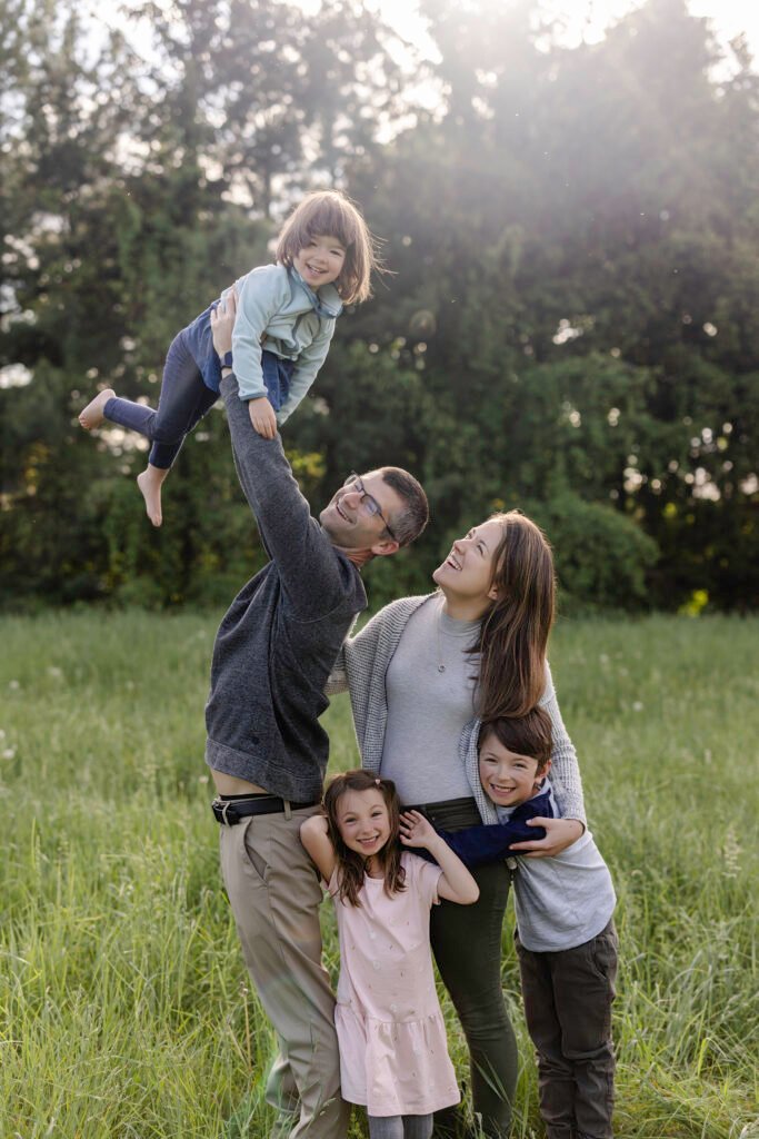 Une famille souriante de cinq personnes se tient dans un champ herbeux. Un adulte soulève un jeune enfant au-dessus de sa tête, tandis qu'un autre adulte étreint deux enfants plus âgés à ses côtés. Des arbres et le soleil sont visibles à l'arrière-plan.