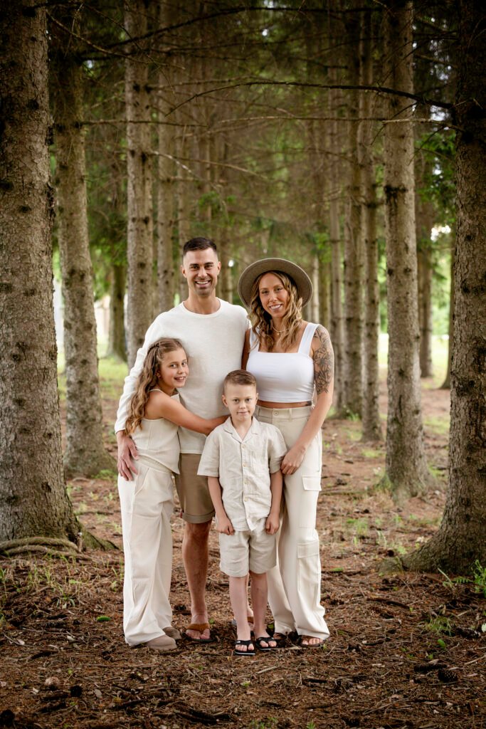 Une famille de quatre personnes se tient debout, proche les uns des autres, au milieu de grands pins. Les adultes sourient derrière deux enfants, tous vêtus de tenues légères et décontractées. La lumière du soleil filtre à travers les arbres à l'arrière-plan.