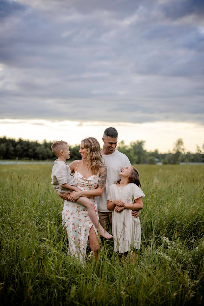 Une famille de quatre personnes se tient dans un champ herbeux sous un ciel nuageux, souriant et riant ensemble. La mère tient un jeune garçon dans ses bras, tandis que le père et la fille se tiennent près d'elle, tous vêtus de vêtements légers et estivaux.