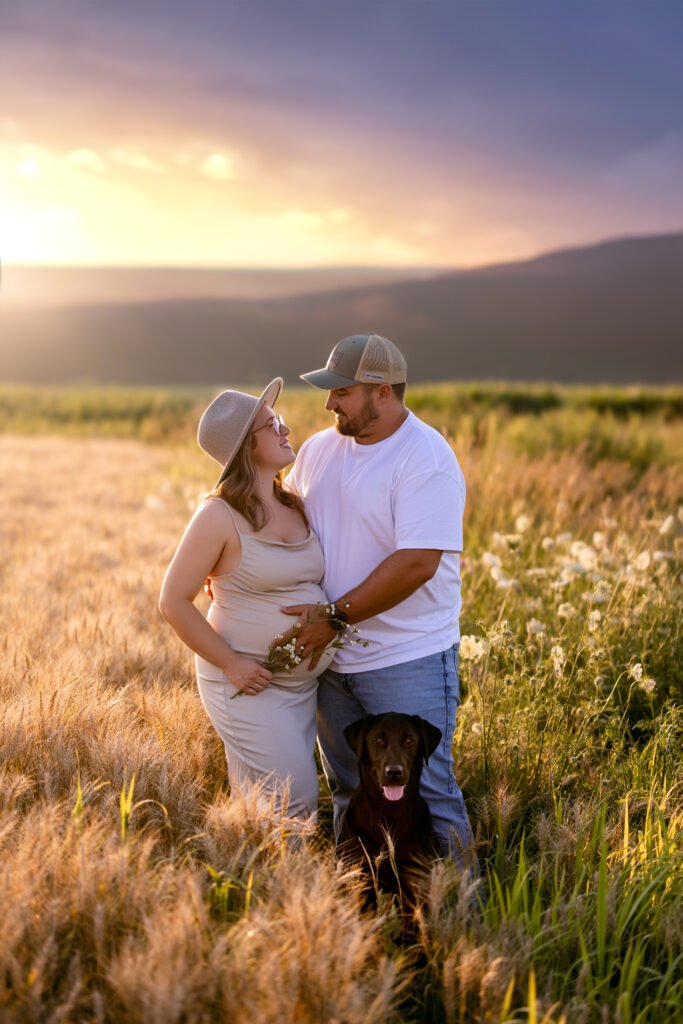 Un couple se tient dans un champ au coucher du soleil, se regardant l'un l'autre. La femme porte un chapeau et une robe légère, tandis que l'homme porte une casquette et un t-shirt blanc. Un chien brun est assis devant eux, au milieu des hautes herbes et des fleurs sauvages.