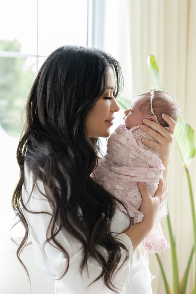 Une femme aux longs cheveux noirs tient un nouveau-né enveloppé dans une couverture en dentelle rose, touchant doucement son front à celui de l'enfant, tandis qu'ils partagent un moment de tendresse près d'une fenêtre éclairée par le soleil.