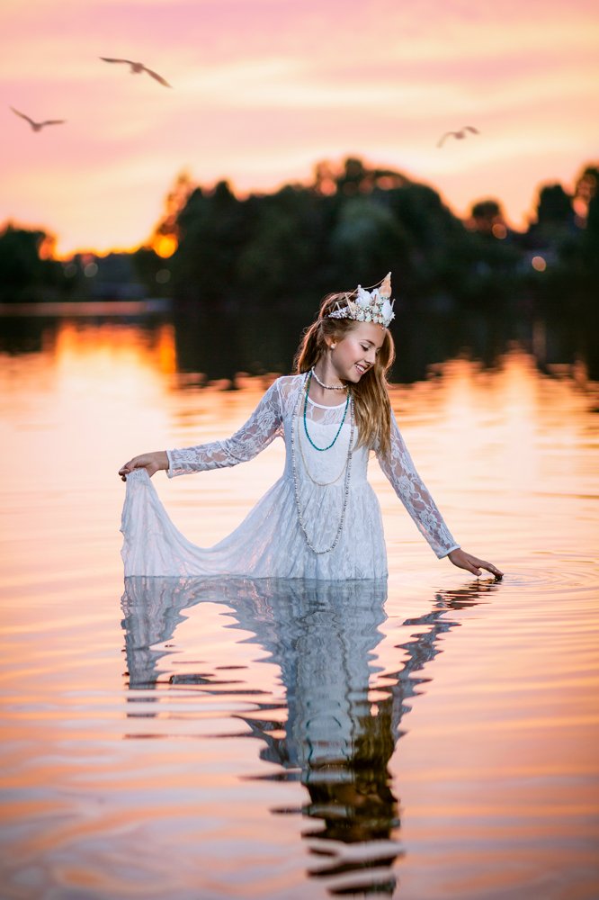 Une jeune fille vêtue d'une robe blanche en dentelle et d'une couronne de coquillages se tient dans un lac au coucher du soleil, en tenant sa robe au-dessus de l'eau. Des arbres et des oiseaux sont à l'arrière-plan, et le ciel s'illumine de teintes roses, orange et violettes.