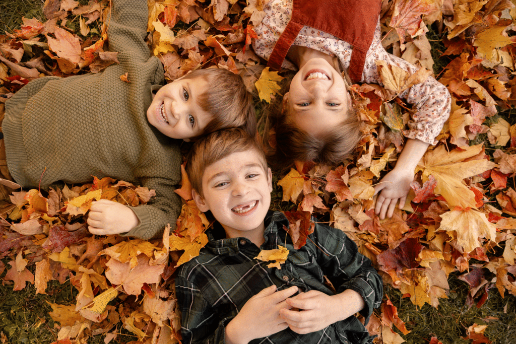 Trois enfants souriants sont allongés côte à côte sur un tas de feuilles d'automne colorées et regardent l'appareil photo. La scène capture un moment joyeux et ludique en plein air, à l'automne.