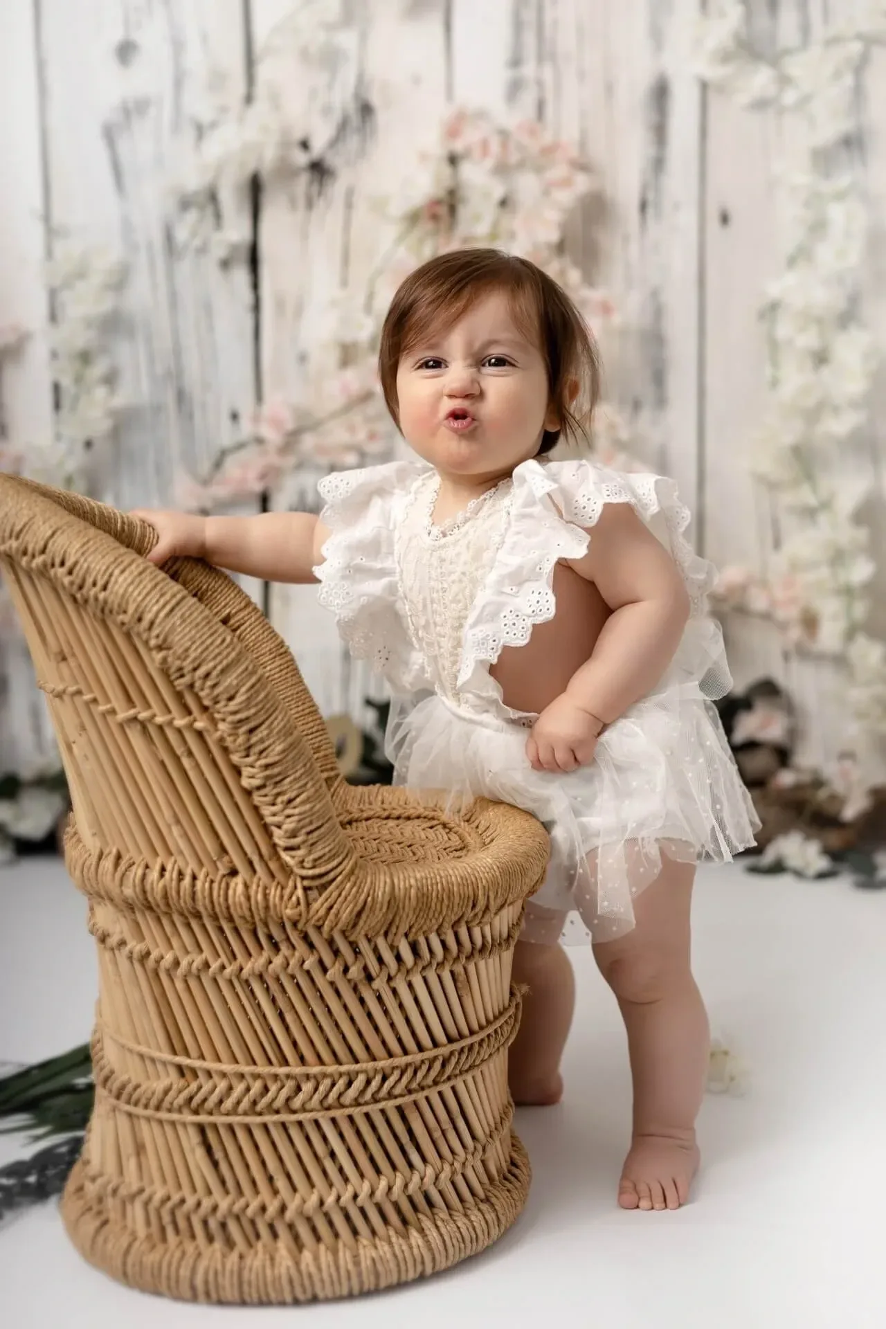 Bambine en robe blanche debout près d’une chaise en osier, dans un décor doux