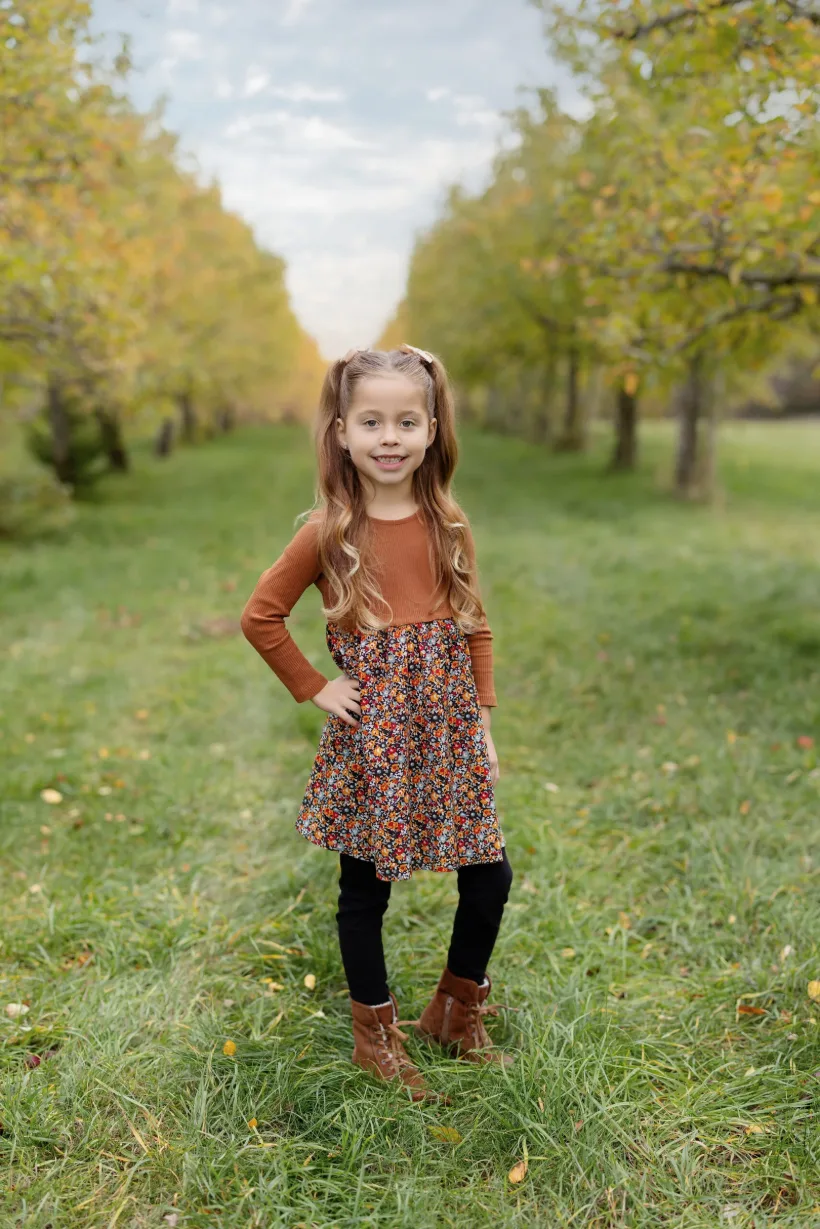 Jeune élève souriante en robe automnale, debout dans une allée d’arbres, portrait scolaire naturel